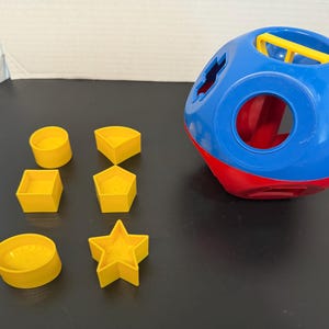 May include: A blue and red shape sorter toy with various cut-out shapes. Includes yellow geometric shapes: cylinder, square, pentagon, semi-circle, and star. The toy is designed for early childhood development and learning.