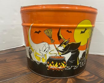 May include: Orange metal Halloween bucket with a wraparound illustration. The design features a witch stirring a cauldron, a ghost, bats, a full moon, and a black cat. The bucket has a white rim and a dark base.