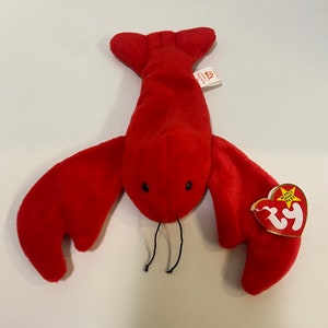 May include: A plush red lobster toy with black bead eyes and black thread antennae. The lobster has large claws and a tail. A red and white heart-shaped tag is attached to the lobster.