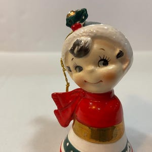 May include: A vintage porcelain Christmas bell ornament featuring a smiling girl wearing a red and white hat and a red scarf. The bell is decorated with green and white stripes and a gold band.