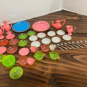 May include: A collection of vintage miniature dollhouse dishes and tea set pieces in various colors. Includes pink teapots, cups, plates, and serving dishes, along with blue and pink serving trays, and small spoons. The set is made of plastic.