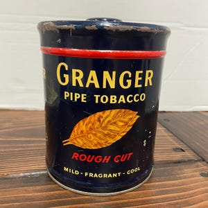 Puede incluir: Lata de tabaco para pipa Granger vintage. La lata cilíndrica es azul marino con una banda roja y letras doradas. El texto dice "GRANGER PIPE TOBACCO" y "ROUGH CUT". También presenta una ilustración de hoja dorada.