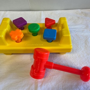 May include: A yellow plastic toy workbench with a red hammer and five colorful shape blocks. The blocks are a purple square, a red triangle, a blue square, a green circle, and an orange plus sign. The workbench has the text "Fisher-Price" printed on it.