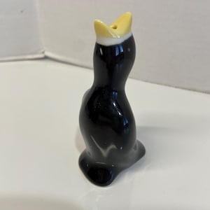 May include: A black ceramic figurine of a bird with a yellow beak and a white band around its neck. The bird is in a seated position with its beak open. The figurine has a glossy finish and is on a white surface.