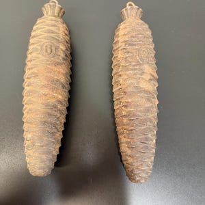 May include: Two rustic, brown pine cone-shaped weights, likely for a cuckoo clock. Each has a textured surface and a small loop at the top. One has the number "6" imprinted on it.
