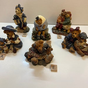 May include: A collection of decorative bear figurines in various poses. The bears are dressed in detailed outfits, some holding objects like teacups and a sailboat. The figurines are on a white surface, with letter tiles scattered around them.