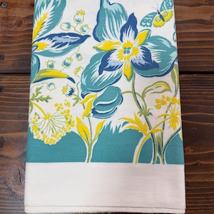 May include: Folded white and teal floral patterned kitchen towel. The design features large blue and yellow flowers, green leaves, and a butterfly. The bottom of the towel has a solid teal band and a white border.