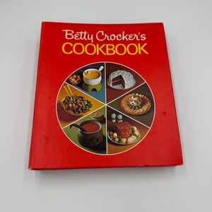 May include: A vintage cookbook titled "Betty Crocker's COOKBOOK" in red with yellow lettering. The cover features a circular design with images of various dishes, including soup, cake, and salad. The book is closed and in good condition.