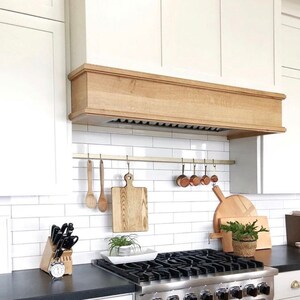 A white kitchen with a black countertop and a stainless steel stove. The stove has a white oven and a black cooktop with a gold utensil rack above with small cutting boards and measuring cups hanging. There is a wooden range hood above the stove. The kitchen has white cabinets and a white tile backsplash. There are wooden cutting boards and utensils hanging on a brass rail above the stove.