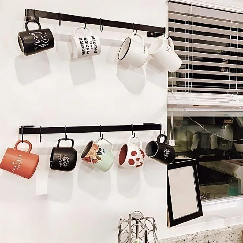 Kitchen with two black metal hook rack mounted on the wall with coffee mugs hanging. There is also a window with shades next to it.