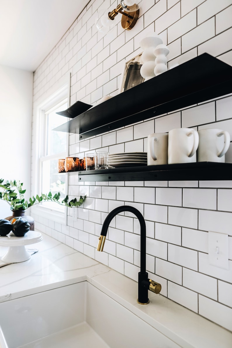 modern kitchen with two metal floating shelves mounted onto a white backsplash. There are dishes and glasses on each black shelf. There is a faucet and wall sconce and plants.