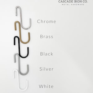 Five S-shaped hooks in different finishes: chrome, brass, black, silver, and white.  The hooks are displayed against a white background with the text "CASCADE IRON CO. METAL HARDWARE" and "www.cascadeironco.com".