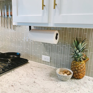 kitchen with under cabinet black paper towel holder mounted under white cabinets. There is sage green tile backsplash and brass cabinet handles.