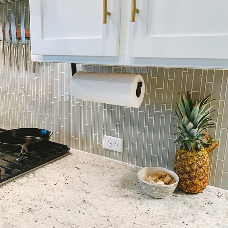 kitchen with under cabinet black paper towel holder mounted under white cabinets. There is sage green tile backsplash and brass cabinet handles.