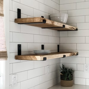 May include: Two wooden shelves with black metal brackets mounted on a white tiled wall. The top shelf has a white bowl on it, and the bottom shelf has a stack of white plates and a potted plant.