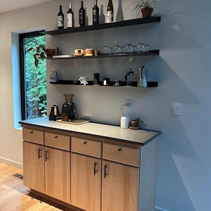 A light wood cabinet with four drawers and two doors. The cabinet has black hardware. Above the cabinet are three black floating metal shelves with various items on them, including wine bottles, glasses, and coffee equipment.