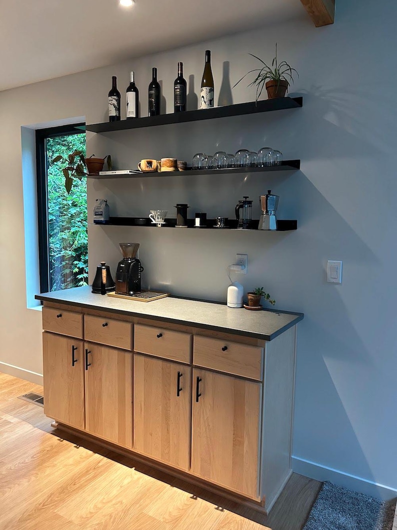A light wood cabinet with four drawers and two doors. The cabinet has black hardware. Above the cabinet are three black floating metal shelves with various items on them, including wine bottles, glasses, and coffee equipment.