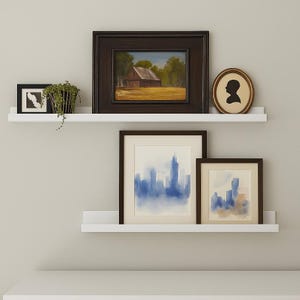Two white floating metal picture ledge shelves with various framed artwork and a mirror. The shelves are mounted on a white wall. The artwork includes a landscape painting, a silhouette of a person, and two abstract paintings. The mirror is a hexagonal shape with a wooden frame.