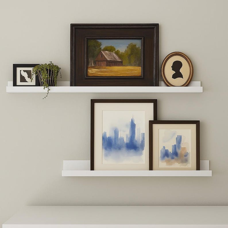 Two white floating metal picture ledge shelves with various framed artwork and a mirror. The shelves are mounted on a white wall. The artwork includes a landscape painting, a silhouette of a person, and two abstract paintings. The mirror is a hexagonal shape with a wooden frame.