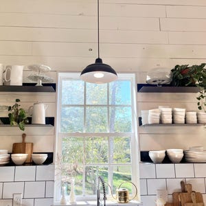 A white kitchen with black metal shelves holding white dishes, a white pitcher, and a glass cake stand. A black pendant light hangs above the window.