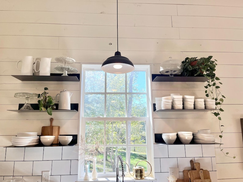 A white kitchen with black metal shelves holding white dishes, a white pitcher, and a glass cake stand. A black pendant light hangs above the window.