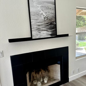May include: A black fireplace with a black mantel shelf and a framed black and white photograph of surfers on a wave above the mantel. The fireplace has a black tile surround and a glass fire screen. There are five white candles inside the fireplace.
