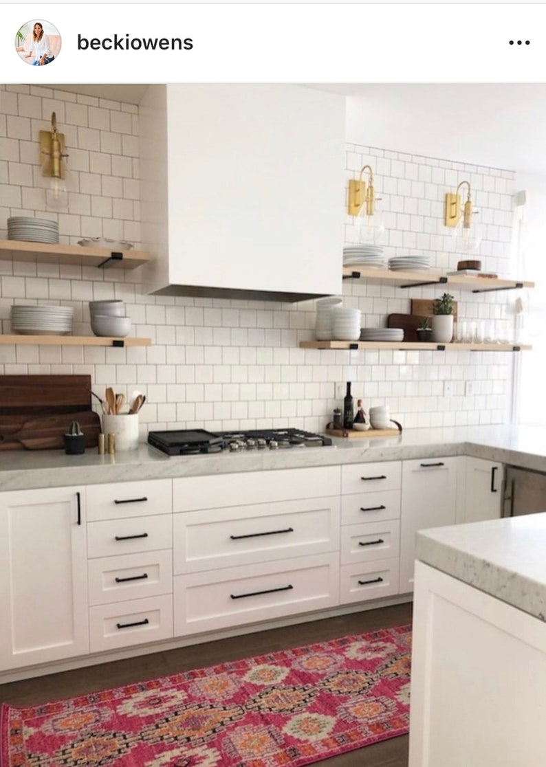 A white kitchen with white cabinets and black hardware. The kitchen has a white subway tile backsplash and a stainless steel stovetop. There are wooden shelves above the stovetop with dishes and plants. The kitchen has a pink and orange patterned rug.