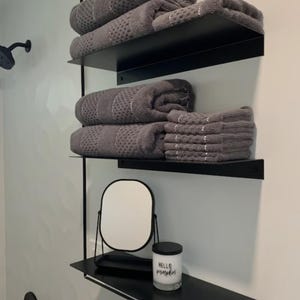 Bathroom with three black floating shelves with gray towels folded on them. The shelves are mounted on a gray wall. The bottom shelf has a small round mirror and a white candle.