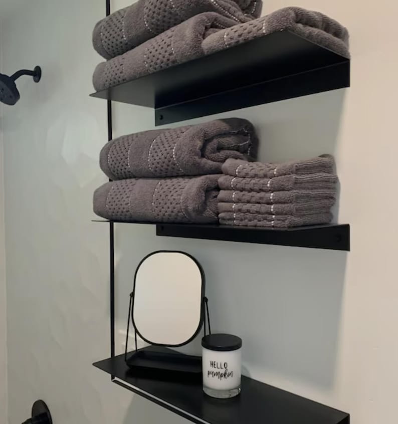 Bathroom with three black floating shelves with gray towels folded on them. The shelves are mounted on a gray wall. The bottom shelf has a small round mirror and a white candle.