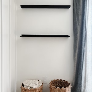 There are two black metal display ledges mounted on a white wall. They are empty and have a front lip. There is a long curtain to the side with two baskets below.