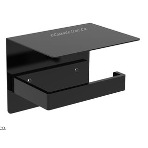 May include: Black metal toilet paper holder with a shelf. The holder has a rectangular shape and a small shelf on top. The text "©Cascade Iron Co." is printed on the shelf.
