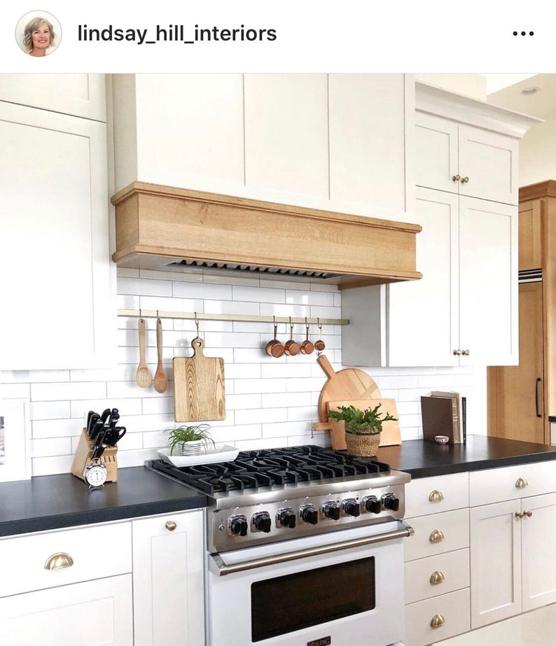A white kitchen with a black countertop and a stainless steel stove. The stove has a white oven and a black cooktop with a gold utensil rack above with small cutting boards and measuring cups hanging. There is a wooden range hood above the stove. The kitchen has white cabinets and a white tile backsplash. There are wooden cutting boards and utensils hanging on a brass rail above the stove.
