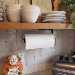 Kitchen with a metal paper towel holder mounted under wood shelf with white dishes. There is ceramic tile backsplash, a toaster, and decor.