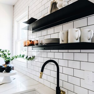May include: A white kitchen with black metal shelves, a white ceramic sink, and a black faucet with a gold handle. The shelves are filled with white mugs, copper mugs, and plates.