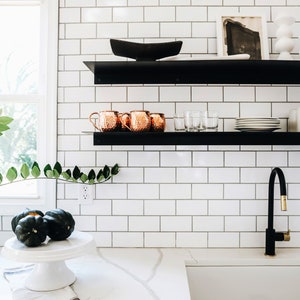 A white tiled kitchen with black floating shelves. The shelves are decorated with copper mugs, clear glasses, white plates, and a black bowl. A brown vase with green leafy branches sits on the counter below the shelves. There is a white countertop with a sink and a black faucet with a gold handle.