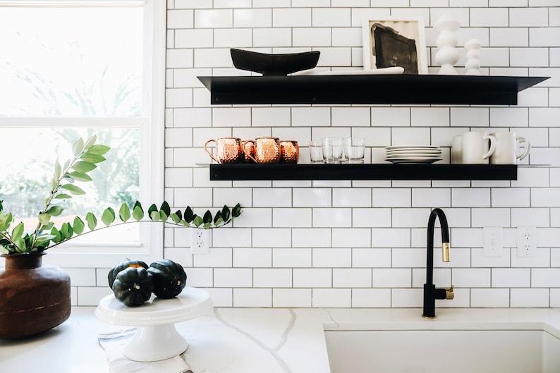 A white tiled kitchen with black floating shelves. The shelves are decorated with copper mugs, clear glasses, white plates, and a black bowl. A brown vase with green leafy branches sits on the counter below the shelves. There is a white countertop with a sink and a black faucet with a gold handle.