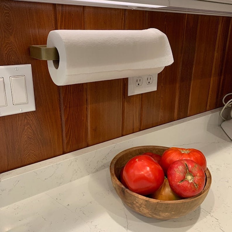 brass  paper towel holder mounted on wood kitchen wall. There is a bowl with fruit on white countertops