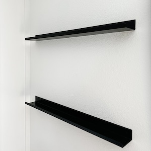 two black metal display ledges mounted on a white wall. They are empty and have a front lip.