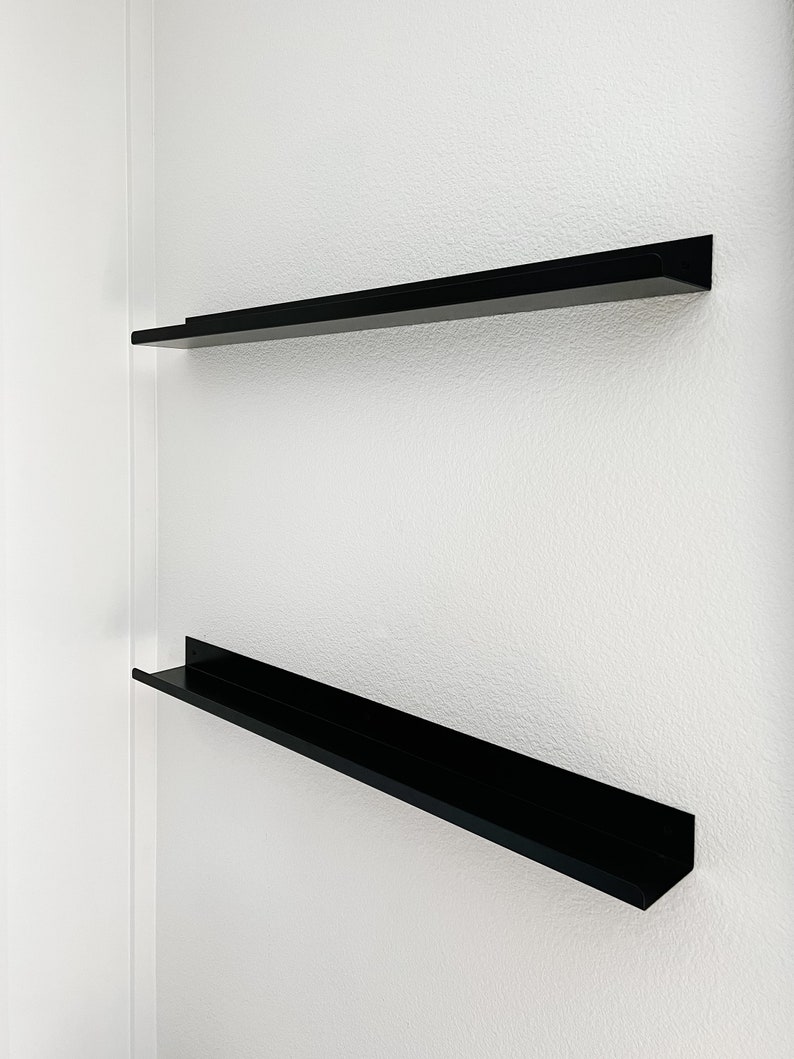 two black metal display ledges mounted on a white wall. They are empty and have a front lip.