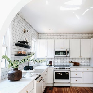 Updated white kitchen with two black metal floating shelves installed on one tiled wall. There are white cabinets, a wood floor, and plants.