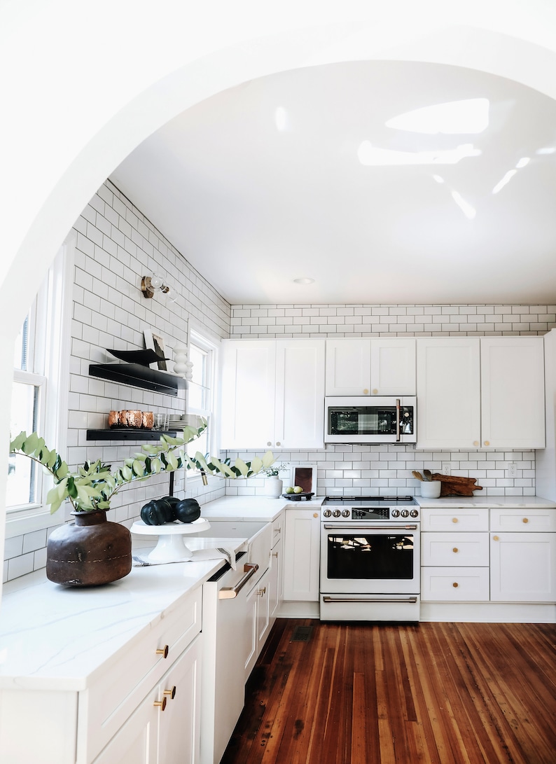 Updated white kitchen with two black metal floating shelves installed on one tiled wall. There are white cabinets, a wood floor, and plants.