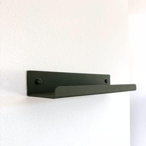 May include: A dark green metal shelf mounted on a white wall. The shelf has a simple, modern design with a flat top and a slightly raised front edge. Two screws secure the shelf to the wall.
