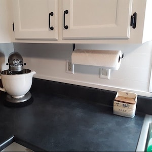 White kitchen with black countertops with black paper towel dispenser holding a paper towel roll mounted under wood cabinet. There is a mixing bowl stand on counter.
