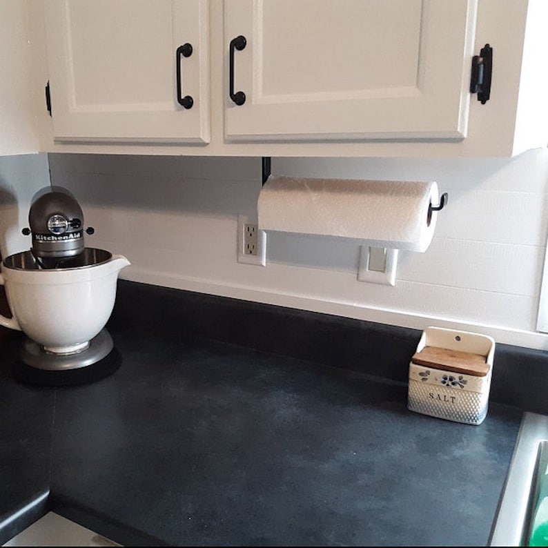 White kitchen with black countertops with black paper towel dispenser holding a paper towel roll mounted under wood cabinet. There is a mixing bowl stand on counter.