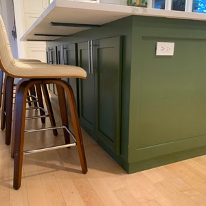 May include: Two light beige bar stools with a wood frame and chrome legs, positioned next to a dark green kitchen island with white countertops and silver cabinet hardware.