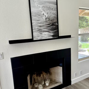 A black fireplace with a black tiled surround and a black floating metal mantel shelf. A black framed photograph of surfers riding waves hangs above the shelf. The fireplace is lit with white candles.