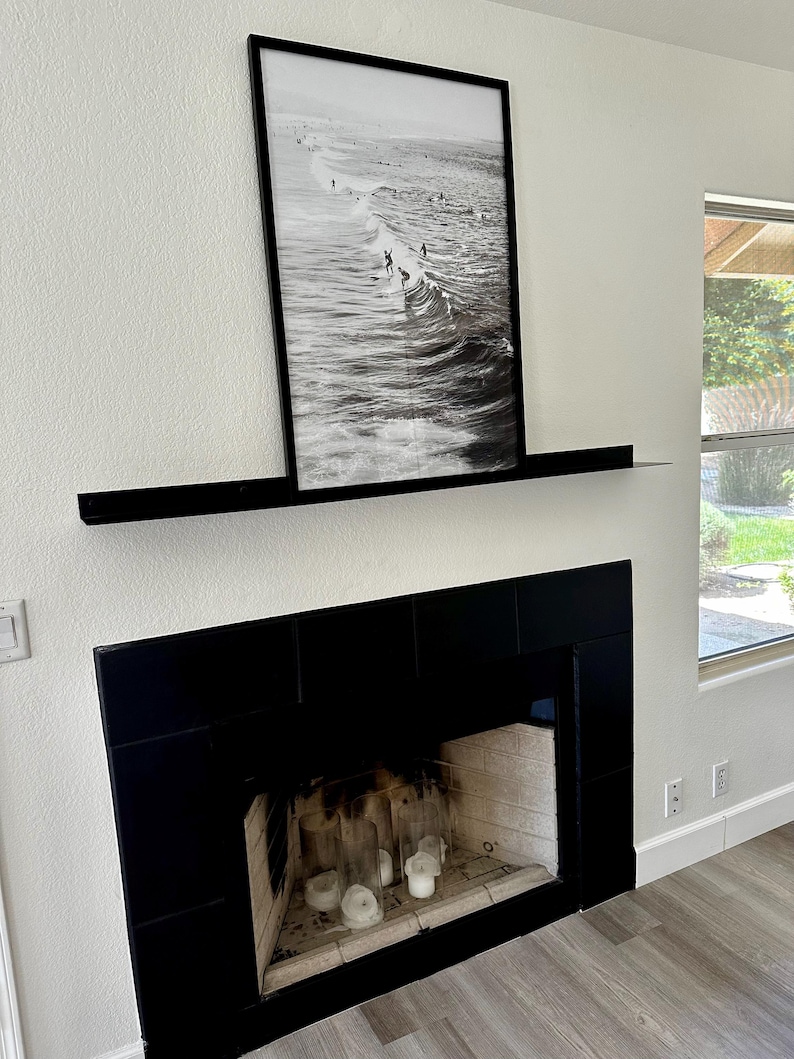 A black fireplace with a black tiled surround and a black floating metal mantel shelf. A black framed photograph of surfers riding waves hangs above the shelf. The fireplace is lit with white candles.