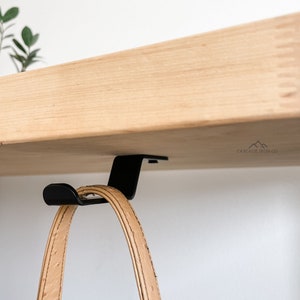 May include: A black metal hook mounted under a wooden shelf, with a tan leather purse hanging from it. The hook is labeled "CASCADE IRON CO."