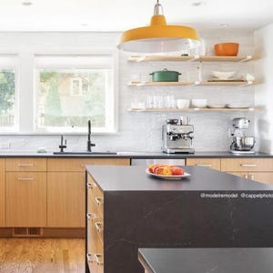 A mid century modern kitchen with light wood cabinets, a black countertop island, and a yellow pendant light hanging above. There are wood floating shelves with white heavy duty brackets. The kitchen features a stainless steel sink, a coffee maker, and a stand mixer.