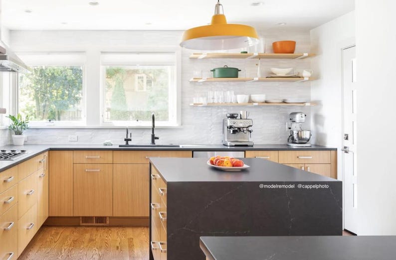 A mid century modern kitchen with light wood cabinets, a black countertop island, and a yellow pendant light hanging above. There are wood floating shelves with white heavy duty brackets. The kitchen features a stainless steel sink, a coffee maker, and a stand mixer.